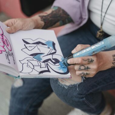 Tattooed hand sketching graffiti art with marker in notebook, showcasing creativity and street style.